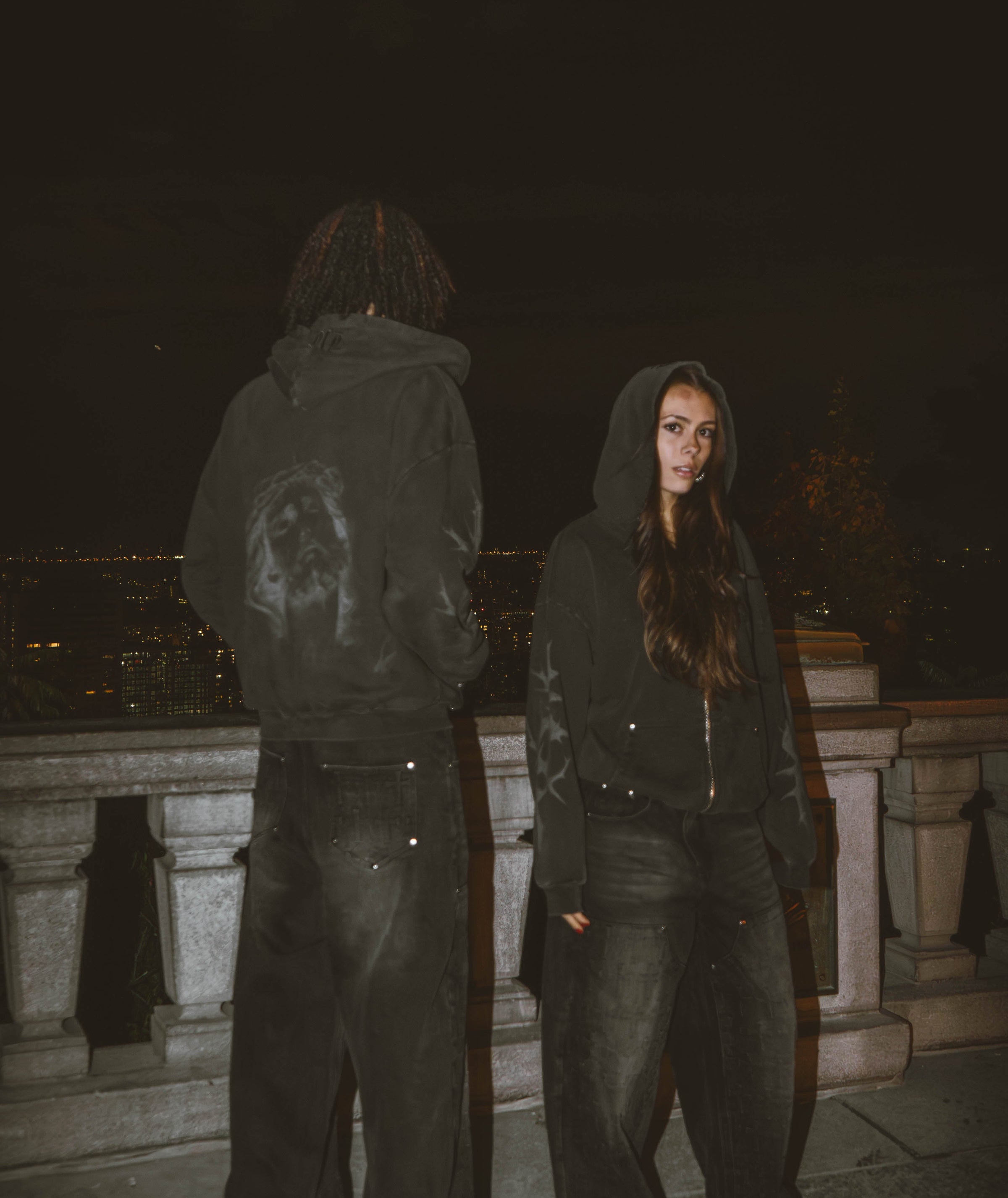 Two people wearing dark hoodies and pants standing on a stone ledge at night.